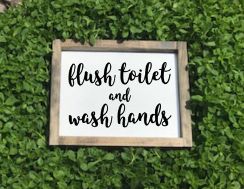May include: A white wooden sign with black text that reads "flush toilet and wash hands". The sign is sitting on a bed of green plants.