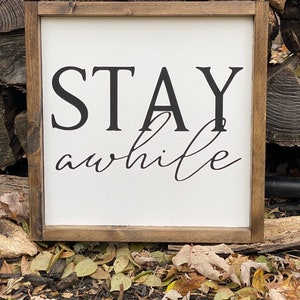 May include: A wooden framed sign with the text "STAY awhile" in black lettering on a white background.