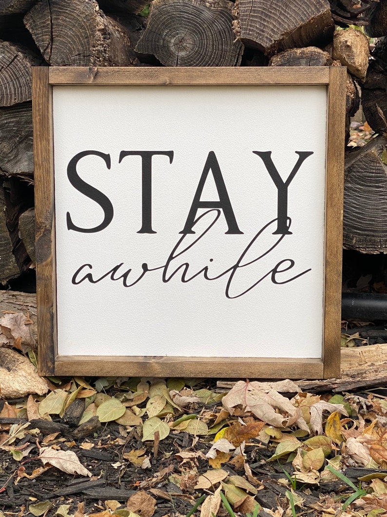May include: A wooden framed sign with the text "STAY awhile" in black lettering on a white background.