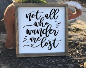 May include: A white wooden sign with a black cursive inscription that reads "Not all who wander are lost".