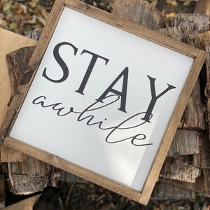May include: A wooden framed sign with white background and black text that reads "STAY awhile". The sign is sitting on a pile of wood.