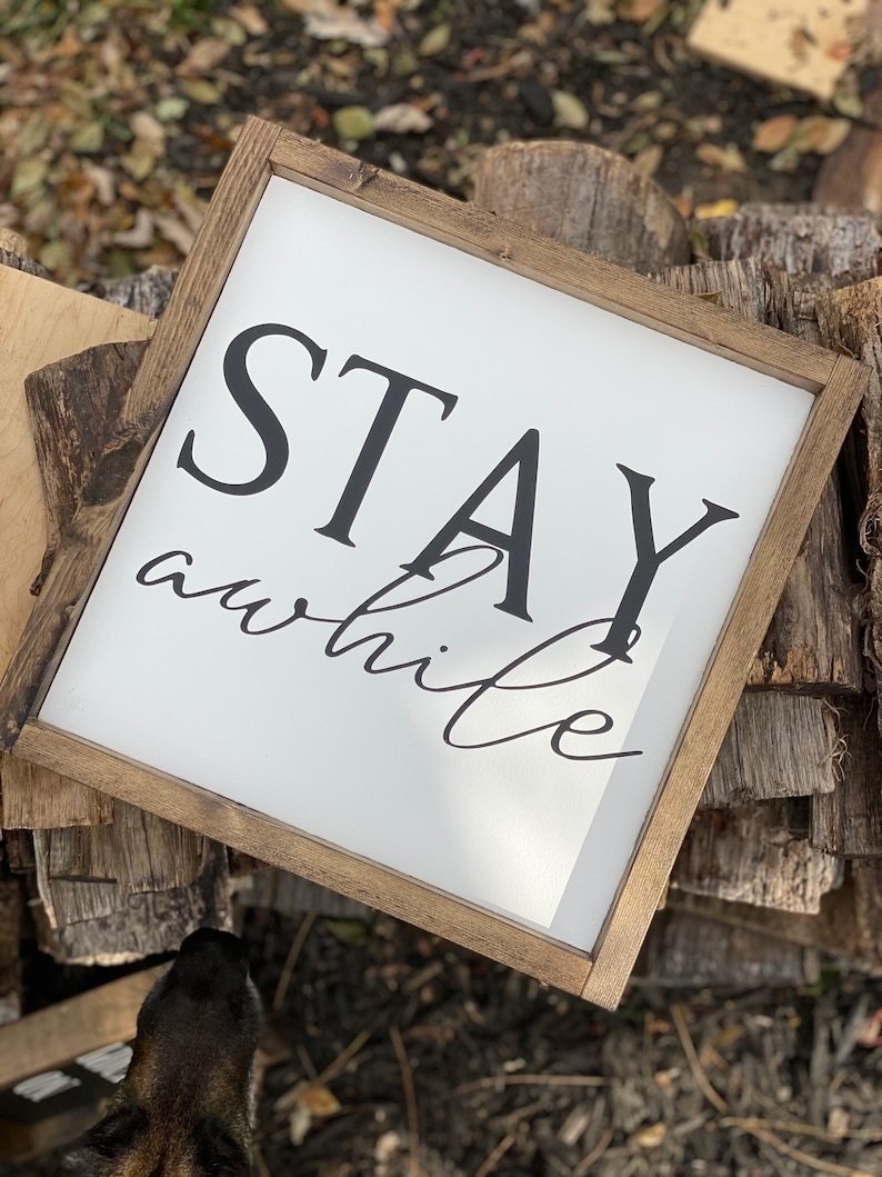 May include: A wooden framed sign with white background and black text that reads "STAY awhile". The sign is sitting on a pile of wood.