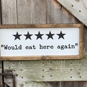 May include: A white wooden sign with a brown frame. The sign has five black stars and the text "Would eat here again".