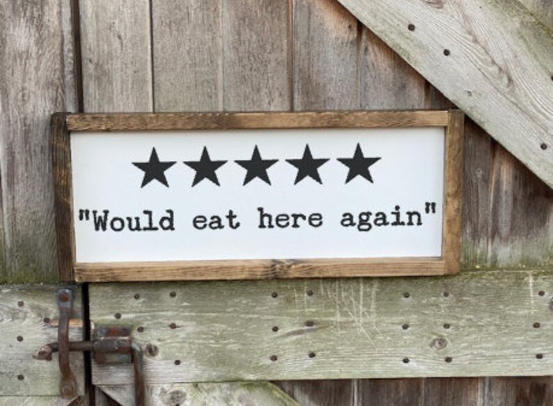 May include: A white wooden sign with a brown frame. The sign has five black stars and the text "Would eat here again".
