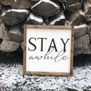 May include: A white wooden sign with a brown frame, featuring the text "STAY awhile" in black lettering. The sign is sitting on a bed of brown wood chips and snow.