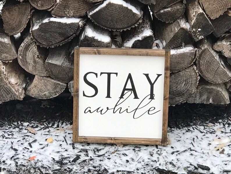 May include: A white wooden sign with a brown frame, featuring the text "STAY awhile" in black lettering. The sign is sitting on a bed of brown wood chips and snow.