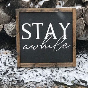 May include: A black wooden sign with white text that reads "STAY awhile". The sign is sitting on a bed of wood chips and snow.
