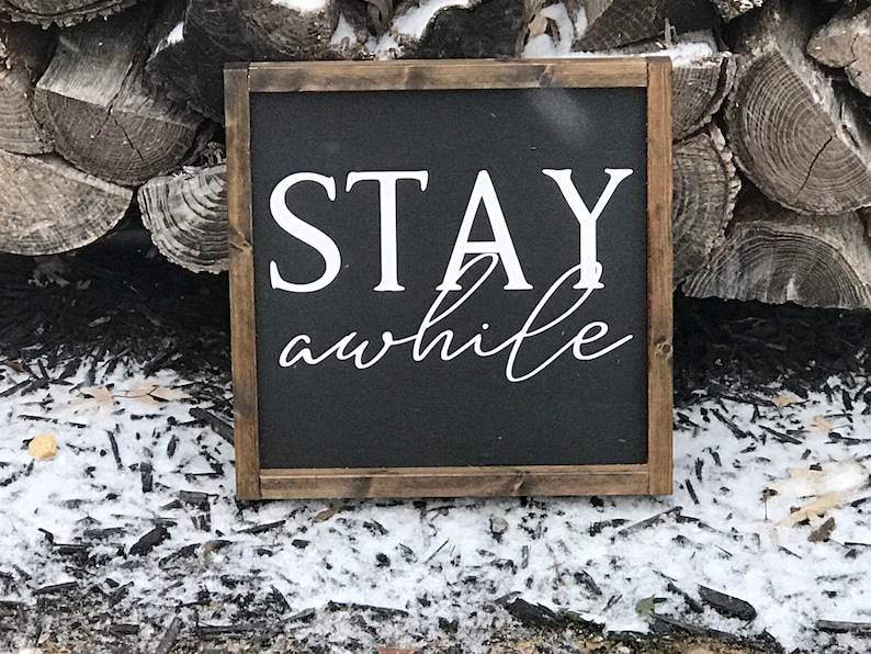 May include: A black wooden sign with white text that reads "STAY awhile". The sign is sitting on a bed of wood chips and snow.