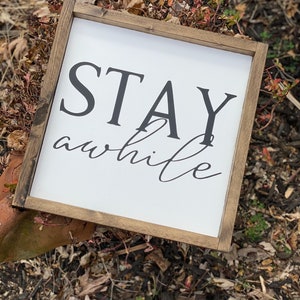 May include: A white wooden sign with a brown frame, featuring the text "STAY awhile" in black lettering. The sign is sitting on a bed of brown mulch.