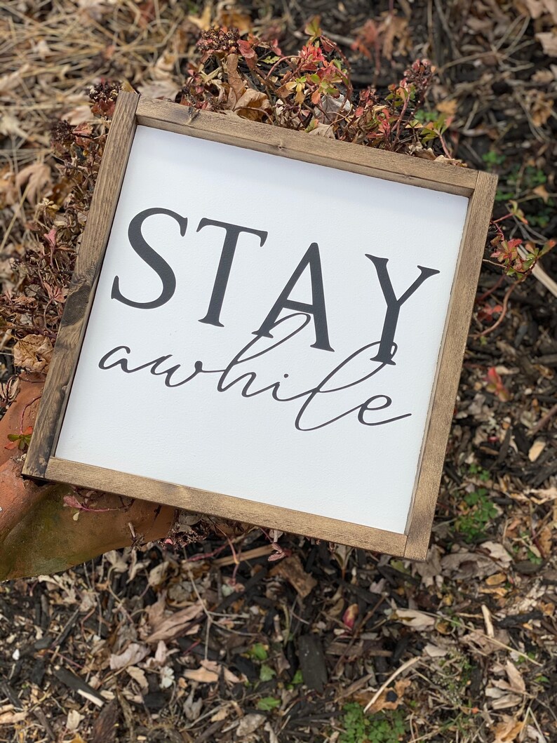 May include: A white wooden sign with a brown frame, featuring the text "STAY awhile" in black lettering. The sign is sitting on a bed of brown mulch.