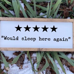 Op de afbeelding: Een wit houten bord met een bruin frame. Het bord heeft vijf zwarte sterren en de tekst "Would sleep here again".