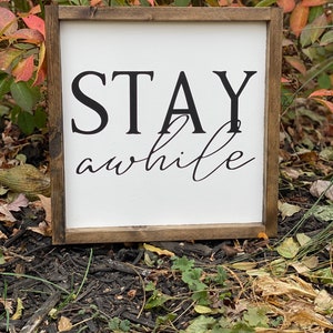 May include: A wooden sign with a white background and black text that reads "STAY awhile". The sign is sitting on a bed of brown mulch.