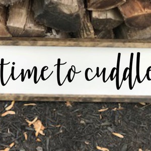 May include: A white wooden sign with a brown wood frame. The sign says "time to cuddle" in black cursive lettering.