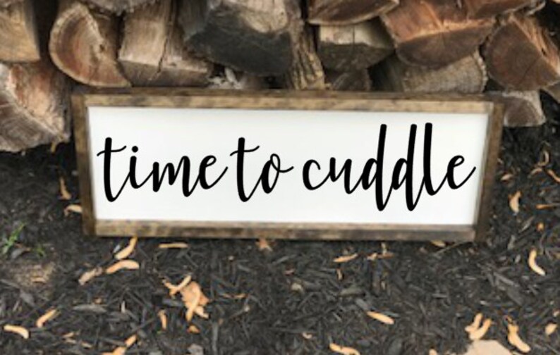 May include: A white wooden sign with a brown wood frame. The sign says "time to cuddle" in black cursive lettering.