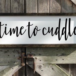 May include: A white wooden sign with black lettering that reads "time to cuddle". The sign is hanging on a wooden wall.