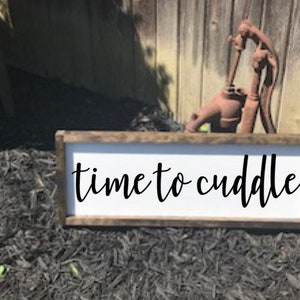 May include: A white wooden sign with a brown frame, the sign reads "time to cuddle" in black cursive lettering.