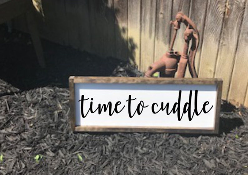 May include: A white wooden sign with a brown frame, the sign reads "time to cuddle" in black cursive lettering.
