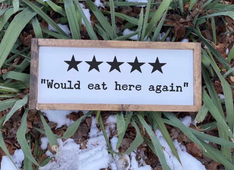 May include: A white wooden sign with a brown frame. The sign has five black stars and the text "Would eat here again".