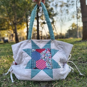 May include: A cream-coloured quilted tote bag with a starburst design. The bag features a patchwork star in shades of blue, red, and green. The handles are striped in green and white. The bag is held up by a hand.