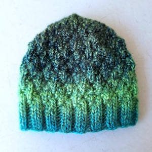 May include: A hand-knitted beanie hat in shades of green and blue. The hat features a ribbed brim that transitions into a textured stitch pattern. The color gradient moves from a bright turquoise at the brim to a deep green at the top.