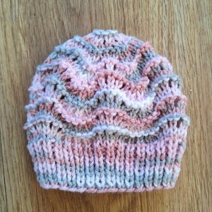 May include: A hand-knitted beanie hat in shades of pink, gray, and white. The hat features a textured, wavy pattern and a ribbed brim. The hat is designed for warmth and style.