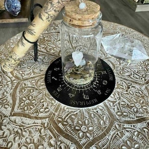 May include: A clear glass jar with a cork lid, containing crystals and small objects, sits on a decorative wooden table. A scroll and a black circular mat with text are also present. The table has an intricate floral pattern.