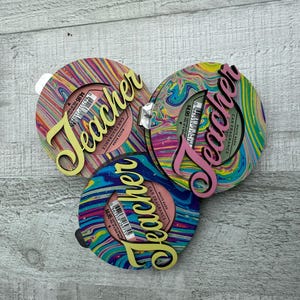 Könnte beinhalten: Drei runde, bunte Holzornamente mit dem Wort "Teacher" in gelben Buchstaben auf jedem. Die Ornamente haben ein marmoriertes, wirbelndes Design in den Farben Rosa, Blau, Grün und Gelb.