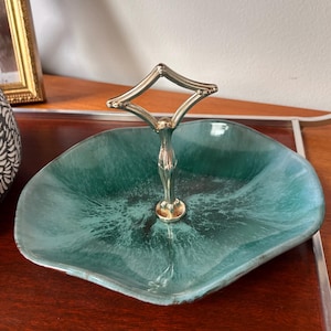 May include: A teal and dark green ceramic dish with an irregular, organic shape and a glossy finish. A central silver-toned metal handle with a diamond-shaped loop at the top is attached to the middle of the dish for carrying or display.