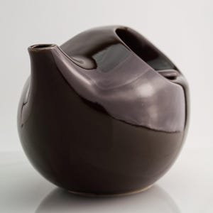 Puede incluir: Una tetera de cerámica esférica de color marrón oscuro con un acabado brillante y reflectante. La tetera presenta un pico corto y curvado que se extiende desde el lateral y una abertura ancha e inclinada en la parte superior, de diseño liso.