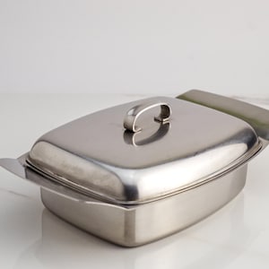 Puede incluir: Una mantequillera rectangular de acero inoxidable con acabado pulido. El recipiente cuenta con una tapa a juego con un asa curva centrada en la parte superior y asas laterales integradas que se extienden hacia afuera desde la base.
