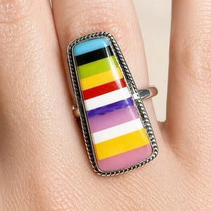 May include: A silver ring with a rectangular, colorful inlay. The inlay features a striped pattern of blue, black, green, yellow, red, white, purple, and pink. The ring has a textured border.