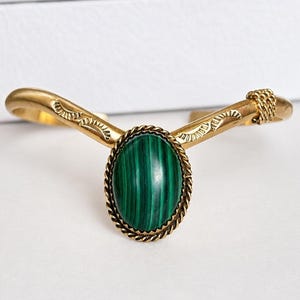 May include: Gold-coloured cuff bracelet with an oval, green malachite stone. The stone is set in a twisted rope-like frame. The bracelet has a V-shape design with etched details and a chain accent on the ends. Jewellery.