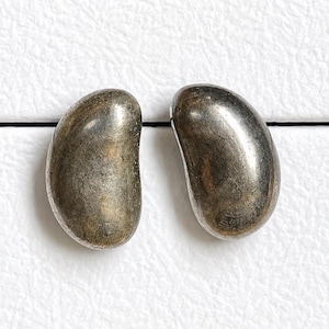 May include: Two silver-toned, bean-shaped earrings. The earrings have a smooth, polished surface with slight variations in color. They are set against a white background.