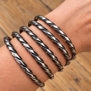 May include: A set of five silver-toned metal bracelets, each featuring a twisted rope design. The bracelets have a polished, reflective finish and are worn stacked together on the wrist, creating a continuous spiraled appearance.