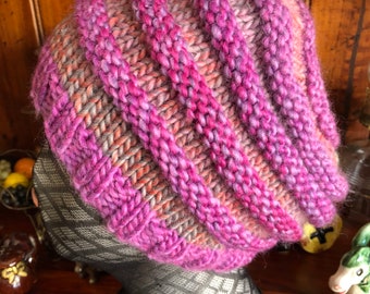 May include: A purple and pink knitted hat with a cable knit pattern. The hat is on a black and grey mannequin head.