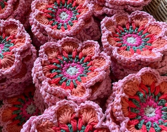 May include: A collection of stacked, handmade crochet coasters in a flower shape. The coasters feature a vibrant color palette of pink, coral, green, and white. The coasters are in a woven basket, showcasing the handmade quality and floral design.