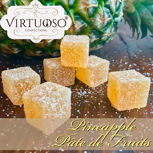 May include: A close-up of six small, square, yellow-colored pineapple pate de fruits candies dusted with white sugar crystals. The candies are arranged on a brown wooden surface. The text "Pineapple Pate de Fruits" is written in a decorative font below the candies.