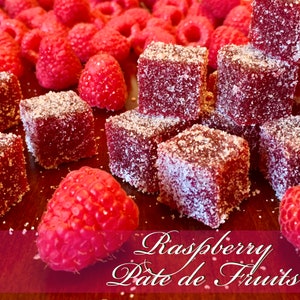 May include: Close-up of raspberry pate de fruits, a type of French fruit jelly candy, dusted with sugar crystals. The candy is arranged on a wooden surface with fresh raspberries scattered around.