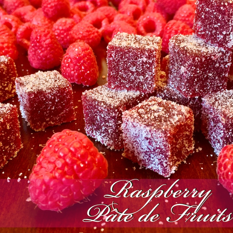 May include: Close-up of raspberry pate de fruits, a type of French fruit jelly candy, dusted with sugar crystals. The candy is arranged on a wooden surface with fresh raspberries scattered around.