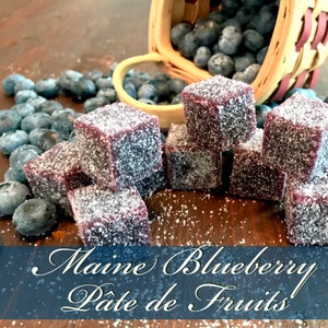 May include: A close-up of a wooden surface with a pile of small, square, purple, sugar-coated candies. The candies are arranged in a pyramid shape. There are blueberries scattered around the candies. The text "Maine Blueberry P&acirc;t&eacute; de Fruits!" is written in a decorative font on a blue background.