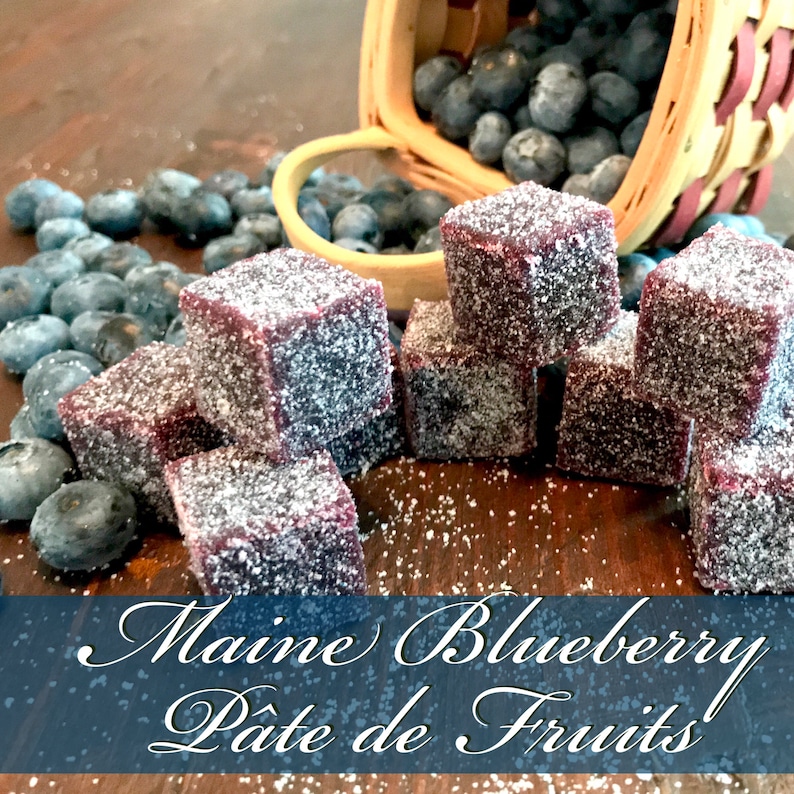 May include: A close-up of a wooden surface with a pile of small, square, purple, sugar-coated candies. The candies are arranged in a pyramid shape. There are blueberries scattered around the candies. The text "Maine Blueberry P&acirc;t&eacute; de Fruits!" is written in a decorative font on a blue background.