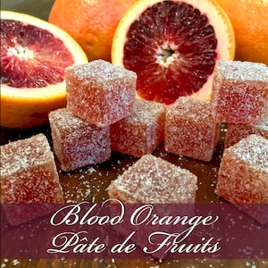 May include: Blood orange p&acirc;te de fruits, a type of French fruit candy, are arranged on a wooden surface with slices of blood oranges in the background. The candy is dusted with sugar crystals.