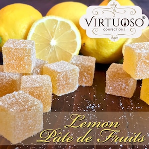 May include: Lemon Pate de Fruits candies from Virtuoso Confections. The candies are square-shaped and dusted with sugar. They are arranged on a wooden surface with lemons in the background.