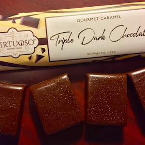 May include: Four dark brown gourmet caramel candies with the text "Triple Dark Chocolate" and "Gourmet Caramel" on a white label with a yellow and black striped background. The label also has the text "Virtuoso Confections" in a brown oval with a gold border.