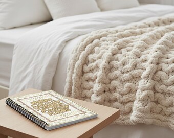 May include: A cream-colored, chunky knit blanket draped over a bed with white linens. A spiral-bound notebook with gold text and floral accents sits on a small wooden table in front of the bed. The notebook reads "Where the snow glows, snow glows, snow glows."