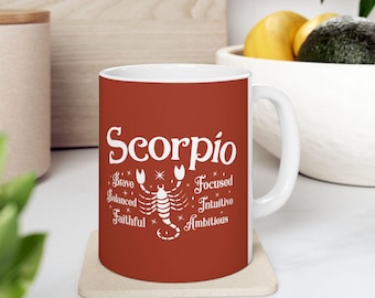 May include: A white ceramic mug with a red background featuring the word "Scorpio" in white script. Below the word is a white scorpion illustration and the words "Brave, Balanced, Faithful, Focused, Intuitive, Ambitious."