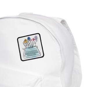 May include: White backpack with a colorful embroidered patch of a typewriter with flowers on top. The patch is surrounded by a black outline.