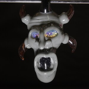 May include: A gray glass pendant with a demonic face, horns, and iridescent eyes. The pendant is hanging from a silver chain.