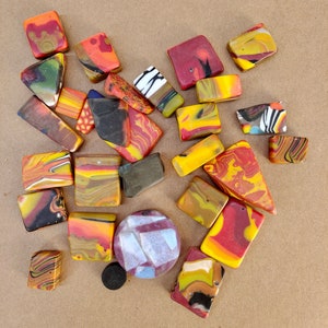 May include: A collection of colourful, abstract, geometric polymer clay tiles in shades of red, orange, yellow, black, and white. The tiles are arranged in a loose, organic pattern on a brown background.