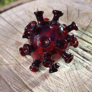 May include: A dark red glass sculpture resembling a virus, featuring numerous rounded protrusions. The spherical object is placed on a wooden surface, showcasing its detailed craftsmanship and unique design.